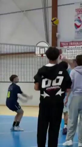 The video shows a group of boys practicing volleyball indoors. They are standing on a blue volleyball court with a net in the middle. One boy is spiking the volleyball over the net. A coach is standing by the net, holding a volleyball and giving instructions to the players. Other boys are standing nearby with volleyballs, waiting for their turn. The room has wooden floors and light walls with wooden trim. The lighting is bright.