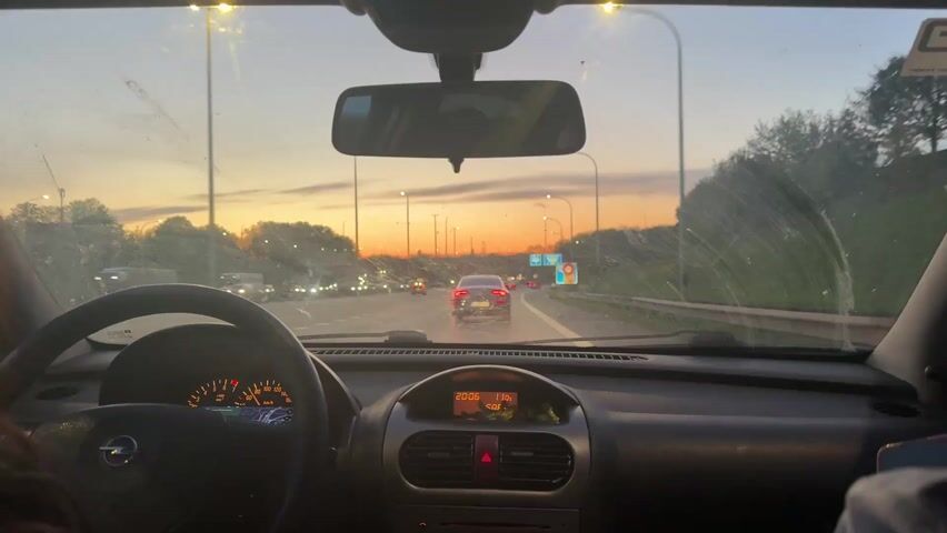 A driver is driving an Opel car down a highway at dusk, with an orange sunset visible in the distance. There are other cars in front and to the left of the driver. The car's dashboard shows the time is 20:06. The windshield is a bit dirty. The car is moving.