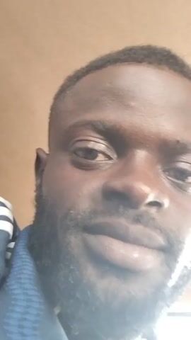A close-up, slightly angled shot of a man's face. The man has dark skin, a short haircut, and a beard. He appears to be looking directly at the camera. He is wearing a blue shirt with white stripes on the shoulder.  The background is a blurred, light brown color. There is a sound of traffic in the background.