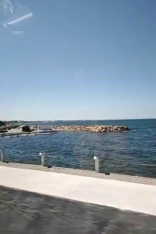 The video captures a view from a moving vehicle, looking out towards a body of water. The water is a deep blue, with visible waves. In the distance, there's a breakwater made of large rocks, and beyond that, more rocky shoreline and some buildings. Several palm trees are visible along the coast. The sky is clear and blue. The foreground shows a concrete barrier and what appears to be the interior of a vehicle, likely a car, with reflections on the windows. The overall scene suggests a coastal drive on a sunny day.