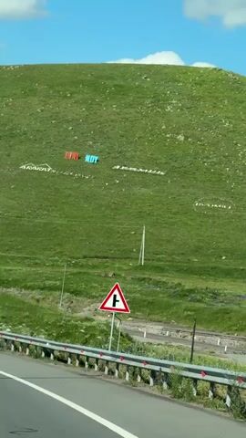 This media file captures a scenic drive in Dzorakap, Armenia. The video pans across a verdant, rolling hillside adorned with large, colorful letters spelling out "HUL NOY" and possibly other words or symbols in white. The hillside is covered in lush green grass, dotted with sparse vegetation and a few trees. Power lines and utility poles are visible on the slope, suggesting a developed area amidst the natural landscape.

In the foreground, a highway runs alongside the hill, bordered by a metal guardrail and a concrete barrier. A triangular road sign with a black symbol indicating a side road junction is prominently displayed near the highway. The sky is a vibrant blue, with fluffy white clouds drifting lazily, indicating a pleasant, sunny day. The time of day appears to be late morning or early afternoon, given the bright sunlight and clear visibility.

The overall impression is one of a journey through a picturesque rural area. The large lettering on the hillside suggests a point of interest, perhaps a landmark or a town name, intended to be seen from a distance. The combination of the natural beauty of the landscape, the man-made elements like the road and signs, and the clear weather creates a sense of peaceful travel and exploration. No people are directly visible in the frame, emphasizing the focus on the scenery and the drive itself. The visible text, "HUL NOY," is a key detail, potentially offering a clue to the specific location or significance of the hillside display.