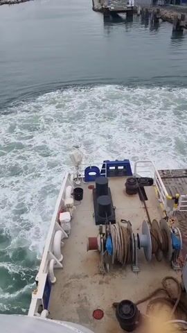 A high-angle static shot captures the stern of a white ferry with a flat deck and various industrial equipment, including coiled ropes and winches. The ferry is moving away from the shore, creating a turbulent white wake in the calm blue-green water. In the background, a small port with docks and buildings is visible, slowly receding as the ferry departs. The sky is overcast and grey.