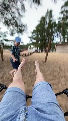 The video shows a first-person perspective of someone sitting in a swing or hammock-like seat. Their legs and denim shorts are visible in the foreground, with their bare feet extended upwards. In the background, on a sandy area surrounded by pine trees, a young child wearing a blue cap and a patterned t-shirt can be seen playing. The child is barefoot and moving around. A hammock is visible in the middle ground, strung between two wooden posts. The scene appears to be outdoors in a park or recreational area, with a cloudy sky overhead. The swing or hammock is in motion, causing the background to blur slightly.