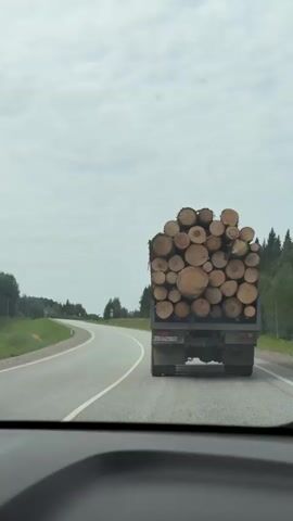 The video shows the rear view of a log truck driving on a highway. The truck is loaded with a large quantity of cut logs. The camera appears to be from inside another vehicle, looking forward through the windshield. The road is paved and appears to be a two-lane highway with a shoulder. On either side of the road are trees and greenery, indicating a rural or forested area. The sky is overcast. The truck has a license plate that is partially visible. The logs are of various sizes and are stacked high on the truck bed. The truck is in motion, driving away from the camera.