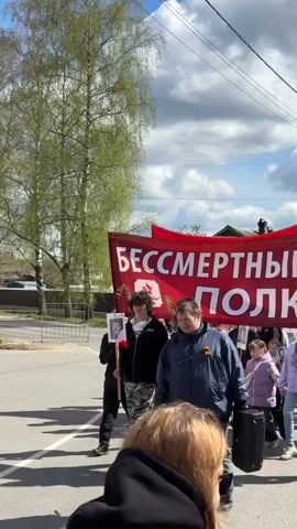 A group of people are marching on a street under a partly cloudy sky. A large red banner reading "BESSSMRTNYI POLK" ("Immortal Regiment") in Russian is held up by several individuals at the front. Many of the people are carrying framed photos, possibly of veterans. Some are wearing orange-and-black ribbons. The environment includes a street, trees, and houses in the background. The group seems to be participating in a commemorative event.