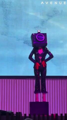 A person in a camera-shaped mask and a sparkly black suit with fringed pant legs dances on a small platform in front of a large screen displaying abstract patterns and the word "AVENUE." The stage is lit with pink and blue lights. The person's hands are gloved in red.