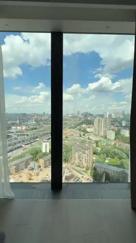 The video presents a sweeping panoramic view of a large city from a high-rise window on a bright, sunny day. The camera slowly pans from right to left, revealing a sprawling urban landscape under a clear blue sky adorned with white, fluffy clouds.

In the immediate foreground, a portion of the window frame and a light-colored floor with a floor-level ventilation grate are visible.

The midground features a dynamic mix of urban elements:
- Two active construction sites are prominent, equipped with multiple cranes, heavy machinery, and ongoing excavation work.
- Numerous multi-story residential buildings, some appearing older, are interspersed with green areas and trees.
- A large, multi-level highway interchange with a significant volume of car traffic is a central feature of the landscape.

As the camera continues to pan to the left, a tall, modern skyscraper with distinctive red accents becomes visible, standing out against the backdrop of other city buildings and the vast highway network.

The background extends into a distant city skyline, showcasing a mix of high-rise buildings and other urban structures that gradually fade into a hazy horizon. No people or visible text are present in the video. The scene depicts a bustling metropolitan area, consistent with a major city like Moscow, which the location context suggests.