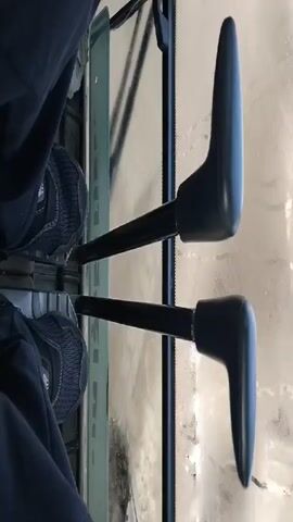 The video shows a person's feet, wearing dark shoes, positioned in front of two black levers. The levers appear to be part of a control system, possibly in a vehicle. The left lever is closer to the viewer and is positioned vertically. The right lever is slightly further away and is angled downwards. Both levers have a dark, rounded handle at the end. The background is blurred and appears to be a reflective surface, possibly a window or a screen, showing indistinct shapes and light. The lighting is subdued, casting shadows on the levers. The overall impression is a close-up view of the controls and the operator's feet in a confined space.