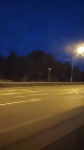 The video shows a view from a vehicle moving along a highway at dusk or night. Streetlights illuminate the road, casting long streaks of light. Trees and a dark sky are visible in the background. A red car passes by in the opposite direction, its motion blurred by speed.