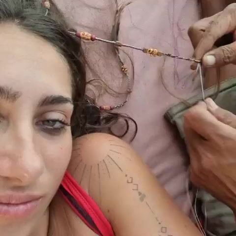 A woman with dark hair and light green eyes looks at the camera as someone makes a beaded hair extension. The person is adding white string to a purple braided extension, which includes various beads, and they are placing the extension on top of the woman's head. The woman is wearing a red top and has two tattoos on her shoulder. One of the tattoos is a rising sun, and the other features various small diamond, triangle, and line shapes.