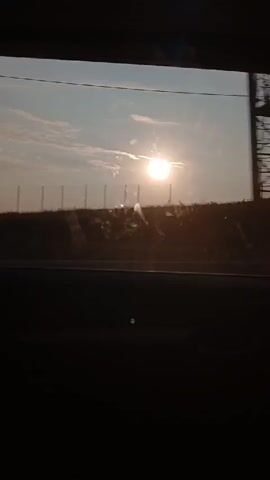 The video shows a view through a car window at sunset. The sun is bright and low on the horizon, casting a warm glow over the landscape. The landscape appears to be a rural area with a hill and some vegetation. The sky is a pale blue with a few wispy clouds. The sun is partially obscured by a thin layer of clouds, creating a hazy effect. The foreground of the video is dark, as it is the interior of the car, with only the reflection of the sun visible on the window. The video captures the moment as the car travels, with the sun appearing to move slightly across the frame. The overall impression is one of a peaceful and serene journey.