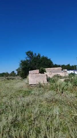The video shows a view of a field with tall grass and some trees. In the foreground, there are some concrete blocks, possibly remnants of a structure. The sky is clear and blue. A dirt path can be seen leading into the distance. A person is walking along the path, and the sound of wind can be heard. The person mentions that there is a nice breeze.