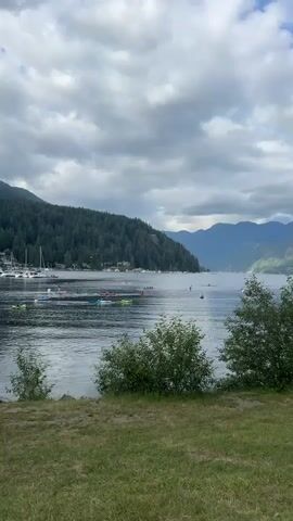 A scenic view of a lake or bay with a forested mountain in the background. The water is calm and reflects the cloudy sky. In the middle ground, several small boats, possibly kayaks or canoes, are scattered across the water. In the far left, a marina with several sailboats is visible. The foreground consists of a grassy area with some scattered bushes. The overall atmosphere is peaceful and natural.