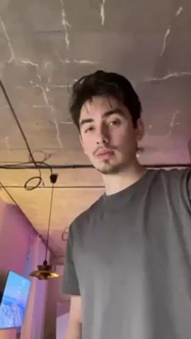 A young man wearing a gray t-shirt stands indoors. The camera looks up at him from below chest level. The man looks down and then looks directly at the camera. The ceiling is concrete with exposed wiring and a bare light bulb. In the background, a television displays a blue sky image, and a lamp with a gold shade hangs down. The lighting in the background is tinged with pink.