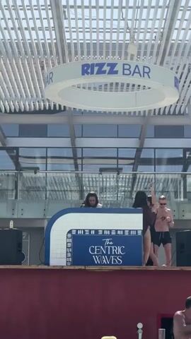 The video shows an outdoor scene at the Rizz Bar, where a DJ is playing music behind a booth marked with "The Centric Waves" logo. A woman in a bikini and sunglasses, also wearing a brown robe, is taking a selfie with a man wearing black swim trunks and sunglasses, who is shirtless. They are both standing on the platform with the DJ.  The Rizz Bar sign hangs above, and the structure has a modern, open-air design with white slats on the roof. A glass-walled building is in the background, and a person can be partially seen in the lower right corner.