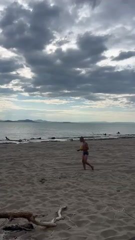 The video shows a sandy beach with a body of water and a cloudy sky in the background. In the foreground, there is driftwood. Two children are visible on the beach. The child on the right is wearing blue swim trunks and holding a yellow object. The child on the left is wearing patterned swim trunks. They appear to be playing together on the beach.