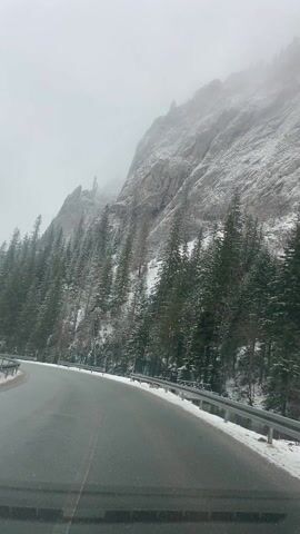 The camera is affixed to the front of a car, providing a driver's-eye view of a winding road in a snowy mountain landscape.  The road is asphalt, wet, and free of snow. To the right, a metal guardrail runs along the edge of the road. Beyond the guardrail, there is a thin layer of snow on the ground. A dense forest of evergreen trees, lightly dusted with snow, rises up the steep mountain slope. The mountain itself is a mix of bare rock and snow. The sky is overcast, and fog obscures the upper reaches of the mountain.  A small building with a green roof is briefly visible on the left. The camera is in motion, driving forward.