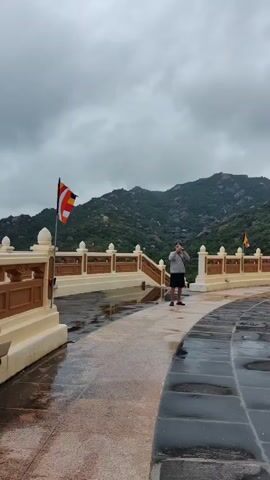 Here's a description of the video:

The video shows a man standing on a platform with a decorative railing. He is taking pictures of a mountain with large rocks on its slopes. The sky is overcast and the ground is wet, possibly from rain. There are flags on poles in the background. The man is wearing a light gray jacket, dark shorts, a blue baseball cap, and sunglasses. He has a dark strap across his body, likely attached to a bag or camera. He holds a smartphone and is focused on capturing an image of the scenery.