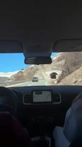 A car is driving towards a tunnel on a sunny day, the car is a Hyundai Venue, the road winds towards the tunnel entrance, there is a passenger in the front seat wearing a white sweatshirt. The car's navigation system is visible in the center console, indicating the route, the mountainside adjacent to the road is partly covered with snow. The driver's left arm is visible, wearing a burgundy sweater. Road signs are visible next to the tunnel entrance.