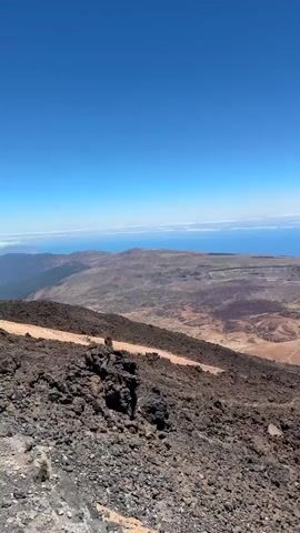 The video pans across a vast, arid landscape under a clear blue sky. In the foreground, rough, dark volcanic rocks cover the ground, with a small wooden railing visible at the bottom right. A dirt path winds through the rocky terrain, marked by a low wire fence. Further into the scene, the landscape opens up to a wide, barren valley with reddish-brown earth and dark patches of what appears to be lava flow. In the mid-ground, a tall metal pylon with cables stretching across it stands prominently, part of a cable car system. Beyond the valley, the vast expanse of the blue ocean stretches to the horizon, meeting the sky. Distant islands or landmasses are faintly visible on the horizon. The overall impression is of a remote, volcanic, and mountainous terrain with a view of the sea. There are no people visible in the video.