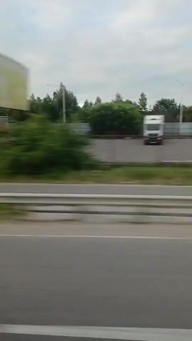 The video shows a yellow truck with a white trailer parked on the side of a road. The truck is viewed from the side, and the video is shot from a moving vehicle. The truck is surrounded by greenery, including trees and bushes. There is a road barrier in the foreground. The weather appears to be overcast. The video is short, lasting only a few seconds, and is blurry due to motion.