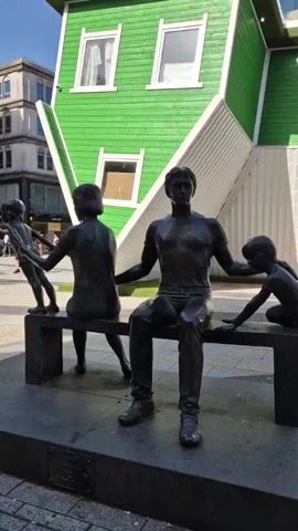 The video shows a bronze statue of a family sitting on a bench. A father sits in the middle, his arms outstretched towards a son on his right and a daughter on his left. Another son stands in front of the bench, holding the hand of the daughter. Behind the statue is a bright green building built upside down. The building has white trim around its windows and a green lawn on top. In the background are several multi-story buildings with various stores. People can be seen walking in the background. The day is sunny.