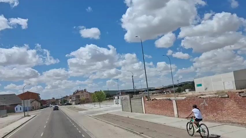 A POV shot from a vehicle moving along a road. The vehicle is on a paved road with white lines dividing lanes. On the left is a sidewalk, a building with a gray roof and brick walls, and a smaller white building. On the right are more buildings, a brick wall, and a large concrete structure that appears to be a silo or industrial building. Beyond this, a castle-like structure sits on a hill. The sky is blue with scattered white clouds. The video shows movement and a passing landscape.
