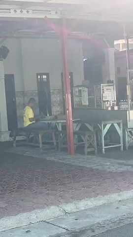 A man in a yellow shirt and blue jeans sits at a picnic table on a tiled patio outside a building. He is looking down at something on the table. To his left is a dark doorway. To his right, a counter with glass displays and menus is visible. The area is outdoors with a red-tiled walkway in the foreground and a concrete curb. The sun is bright and casts a lens flare across the upper part of the image.