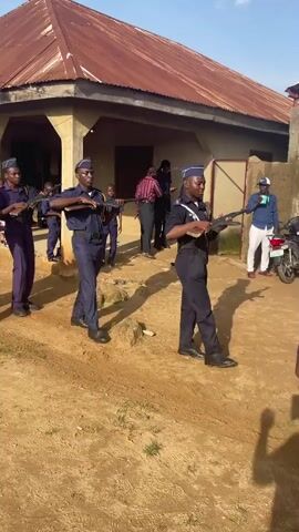 A group of young people dressed in matching dark blue uniforms and caps are marching in formation, holding rifles. They are moving from left to right across the frame. The person in the foreground on the right is partially visible, also in uniform. In the background, several adults are observing the group, including one person in a pink shirt and another in a blue shirt. There is a building with a corrugated metal roof behind the marchers, and a dirt path in front of them. A motorcycle is parked to the right of the frame. The sky is visible and appears to be daytime.