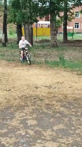 A young boy is riding a bicycle outdoors. He is wearing a white t-shirt and green shorts. The bicycle is black and white. The boy is in focus and appears to be moving towards the camera. The background is blurred, but appears to be a park or grassy area with trees. The sun is out and the lighting is bright. The boy is looking towards the camera and appears to be smiling.