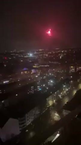 The video shows a nighttime view of a city with sporadic fireworks displays. The scene is taken from a high vantage point, overlooking a landscape of buildings, streets, and scattered lights. Fireworks explode in the distance, creating bursts of light against the dark sky. The video seems handheld.