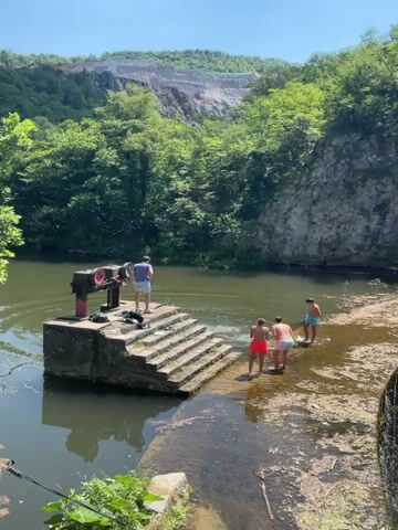 The video captures a bright, sunny day with clear skies at a scenic natural location near Uzice, Serbia. The focal point is a body of water, possibly a river or a lake, with a concrete structure featuring a set of stairs leading into the water. On top of this structure, an industrial-looking piece of machinery, red and metal, is visible.

Several young men are present, enjoying the outdoors. One individual stands on the concrete platform near the machinery, looking out at the water. Two others are in the shallow water near the stairs, and a third is further out in the deeper part of the water. Their attire suggests a casual, recreational setting, likely swimming or enjoying a day out.

The surrounding environment is lush and verdant, with dense green trees covering the hillsides that rise dramatically from the water's edge. A significant geological feature, a large quarry or a rock face with visible strata, dominates the background, adding a unique and striking element to the landscape. The water appears calm with some ripples indicating movement, and the sun casts bright reflections on its surface. The overall atmosphere is one of leisure and outdoor enjoyment in a picturesque, somewhat rugged natural setting. There is no visible text in the video.