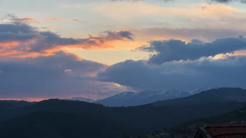The video displays a picturesque sunset over a mountainous landscape. In the foreground, dark, rolling hills are silhouetted against the colorful sky. Above the hills, a dramatic sky showcases a mix of light blue, orange, and purple hues. Dark, heavy clouds are interspersed with patches of fiery orange where the sun's rays break through. In the distance, a range of mountains is visible, their peaks lightly dusted with snow. The light suggests either dawn or dusk. The overall mood is serene and scenic.