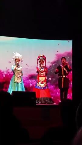 A video shows three people on a stage in front of a large screen displaying a pink and white floral image. On the left, a person in a long, blue, ornate dress and a tall headdress sings into a microphone. In the center, a child or smaller person in a red, ornate dress and a red headdress sings into a microphone. On the right, a man in a dark shirt with colorful accents sings into a microphone. The lighting is dim, with the stage brightly lit and the audience in darkness.