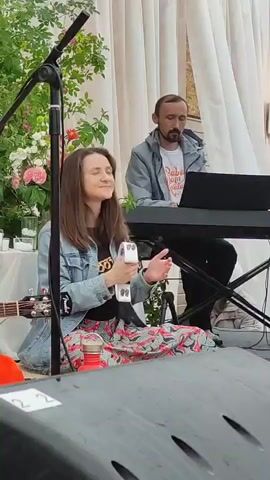 A woman sits cross-legged on the floor in front of a microphone and plays a tambourine. She wears a denim jacket over a t-shirt and a colorful patterned skirt. Behind her, a man sits at a keyboard and looks at a laptop. He wears a grey jacket over a t-shirt. There are flowers and draped fabric in the background. The woman is singing and her eyes are closed.