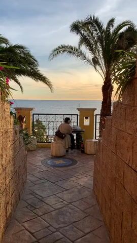 The video shows a sunset over the ocean. There are palm trees in the foreground, and a person is sitting at a table. The person is wearing a light-colored shirt and pants. The table is round and made of wood. There is a small stool next to the table. The ground is paved with stones. There are also some flowers in the foreground. The sun is setting over the ocean, and the sky is orange and pink.