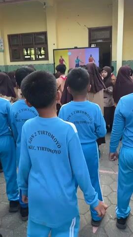 The video shows a group of young children, likely students, standing outdoors in what appears to be a schoolyard. The children are mostly facing a large screen or monitor displaying an image or video of people in colorful outfits. Some children are wearing blue long-sleeved shirts with text that reads "SDN 2 TIRTOMOYO KEC. TIRTOMOYO" on the back. Others are in tan and brown uniforms, possibly scout uniforms. The environment is an open-air area with a building in the background, featuring yellow walls and brown trim. The ground is paved with interlocking gray bricks. One child in the foreground, wearing a blue shirt, turns his head and smiles towards the camera. Another child in a scout uniform on the left side of the frame also appears to be looking in a different direction. The overall atmosphere seems to be of an organized activity or lesson taking place outdoors.