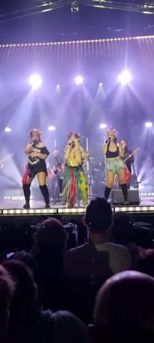 The video shows a musical performance on a brightly lit stage. Three women are positioned in the foreground, singing into microphones. They are dressed in distinctive outfits, including floral headpieces, and appear to be part of a band. Two of the women are wearing shorts and crop tops, while the third wears a black t-shirt dress and tall boots. Behind them, other band members can be seen playing instruments, including drums and guitars. The stage is adorned with numerous spotlights that cast beams of light onto the performers and into the audience. The audience members are visible in the lower portion of the frame, their backs turned to the camera as they watch the performance. The overall atmosphere is energetic and engaging, with the lighting and stage setup suggesting a professional concert environment.
