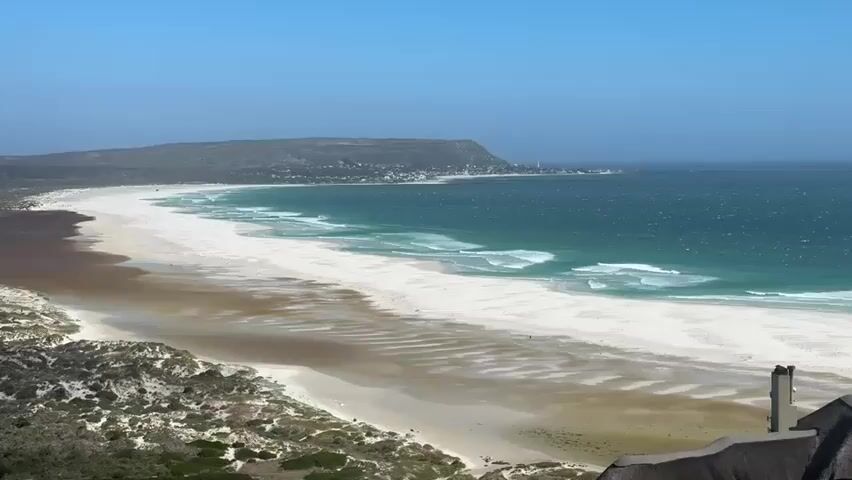 The video captures a panoramic view of a vast, sandy beach with a calm, blue ocean on a clear, sunny day. The beach is characterized by its expansive white sand, transitioning to muddy sand closer to the dunes. Gentle waves break along the shoreline, creating white foamy edges. In the background, a distant headland juts out into the sea, with a small settlement visible on its slopes. The foreground features lush green vegetation and a partially visible thatched roof structure, suggesting a vantage point overlooking the beach. The overall atmosphere is serene and tranquil, emphasizing the natural beauty of the coastal landscape.