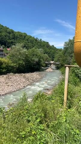 This video captures a scenic daytime view of a river flowing through a valley. The river, with its light greenish-blue water, occupies the center of the frame, bordered by a wide, rocky riverbed. Lush green hills covered in dense trees rise on both sides of the valley, meeting a clear, bright blue sky with a few faint clouds. In the mid-ground, a bridge with a concrete or stone structure spans the river, connecting the two sides of the valley. On the left side, partially obscured by trees, several buildings with white walls and dark roofs are visible on the hillside. On the right side of the foreground, a yellow pipe or post with a wooden cross-piece is visible. The scene is bright and sunny, indicative of good weather. The video is static, showcasing the natural beauty of the area. No people or visible text are present. The geographical context suggests this is a landscape in Krayevsko-Armyanskoye, Russia.