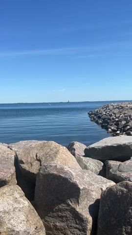 The video shows a calm body of water, likely a bay or ocean, under a clear blue sky with a few scattered clouds. The foreground is dominated by large, grey rocks that form a barrier or breakwater. The water is a deep blue color with gentle ripples on the surface. In the distance, there is a faint silhouette of what appears to be a structure or landmass on the horizon. Several birds fly by. The overall scene conveys a sense of tranquility and natural beauty.