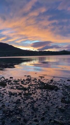 A time-lapse video shows a sunset over a body of water. The sky is filled with clouds in shades of pink, orange, and blue. The water reflects the colors of the sky, and the foreground is a rocky shore. The video is shot from a low angle, looking out over the water. There are no people or animals visible in the video. The environment appears to be a natural landscape, possibly a coastal area.