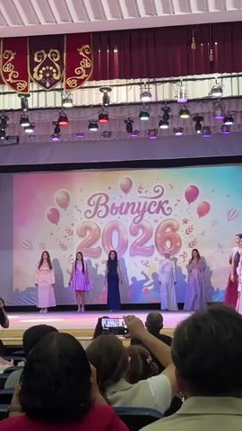 This video shows a graduation ceremony for the class of 2026 at what appears to be a school in Starobaltachevo, Russia. The stage is decorated with a large screen displaying "Выпуск 2026" (Graduation 2026) with colorful balloons and confetti. Six young women, dressed in formal gowns, are on stage, possibly in the process of a performance or receiving recognition. They are spaced out, with some holding hands, and appear to be posing for the audience. The lighting suggests an indoor event, possibly in an auditorium or hall, with spotlights directed at the stage. In the foreground, an audience is seated, with some individuals holding up phones to record the event. The overall atmosphere seems celebratory and formal.
