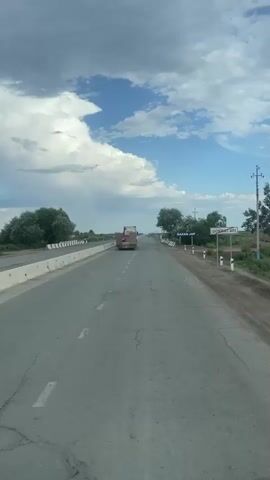 This media file captures a drive along a highway in Qtay, Uzbekistan. The scene is an open, rural landscape under a dynamic sky. The road ahead is paved and shows signs of wear, with visible cracks. A concrete barrier lines the left side of the road, separating it from a grassy verge and some trees. On the right side, a white picket fence with black and white striped posts borders the road.

The primary object on the road is a large truck, appearing to be a flatbed with cargo, driving away from the camera. Several other vehicles, including what appears to be a white car and a motorcycle, are also visible in the distance, suggesting ongoing traffic. A few trees are scattered throughout the landscape, and in the far distance, the horizon is flat and hazy.

The sky is a prominent feature, filled with a mix of bright blue sky and large, dramatic white clouds that suggest either impending rain or recent passing showers. The sunlight filtering through the clouds creates a bright, daytime scene.

Two signs are visible on the right side of the road. The closer sign has blue text on a white background, and appears to read "QAZAQ JAP". Further down the road, another blue sign with white text is visible, though the text is too small to be clearly discerned.

The overall mood is one of travel and open space. The vast sky and the straight road evoke a sense of journey. The weather appears to be fair but with active cloud formations, adding a touch of drama to the otherwise serene setting. The time of day appears to be late morning or early afternoon, given the brightness of the light.