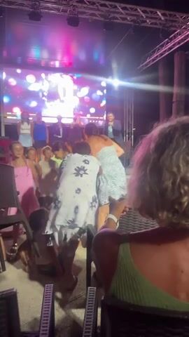 The video shows an outdoor evening event with a stage set up. On the stage, a man in a suit and tie is speaking into a microphone, and a woman in a patterned dress is standing next to him, facing him. There are other people on the stage behind them, including children and adults. A large screen behind them displays a colorful, abstract pattern of bokeh lights. In the foreground, the backs of several audience members are visible, with one person in a green top in sharp focus. The atmosphere appears to be festive, with bright stage lighting illuminating the scene.
