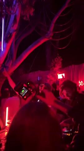 The video depicts a crowded outdoor gathering, possibly a party or event, at night. The scene is illuminated primarily by red and blue lights. A DJ booth with visible equipment and people operating it is in the foreground. Several people are visible, many with their arms raised, possibly dancing or gesturing to the music. Some individuals are holding phones, likely recording or taking pictures. The background includes tree branches and foliage. The overall ambiance suggests a vibrant and lively atmosphere.
