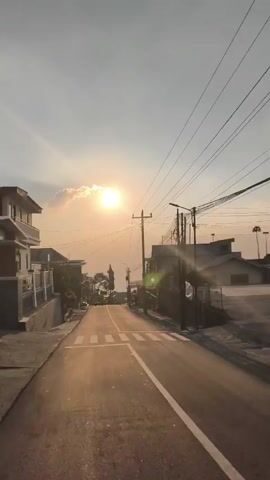 A vertical video shows a paved road leading uphill towards a bright sun breaking through clouds. On either side of the road are houses and utility poles with many wires. The sun is casting a golden glow over the scene. The road has white lane markings and a crosswalk. The sky is a pale orange and white. The overall impression is a serene, possibly rural, setting at sunset or sunrise.