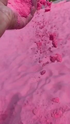 A hand holding pink powder pours it onto a larger pile of the same pink powder, which appears to be sand. The powder is fine and powdery, and it creates a cloud of dust as it falls. The scene is lit by natural light, and the background is out of focus. There are some subtle shimmering particles visible within the powder, suggesting it may contain glitter or a similar reflective material.