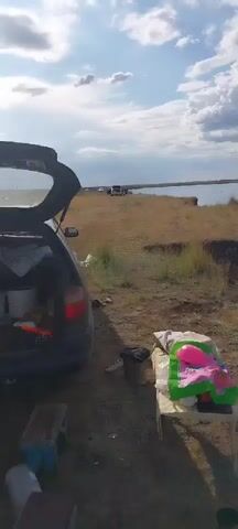 The video pans across a campsite next to a large body of water. In the foreground, a car trunk is open, revealing various items including buckets and bags. A makeshift bed or seating area is set up with blankets, a green mat, and children's toys, including a pink ball. A small blue stool is also visible. The landscape consists of dry grass and dirt, with a steep, eroded bank leading down to the water. In the distance, several cars are parked on a grassy area overlooking the water. The sky is partly cloudy with dramatic cloud formations. A person is seen sitting on the edge of the bank in the middle ground, facing the water. The overall impression is a casual outdoor gathering or picnic.