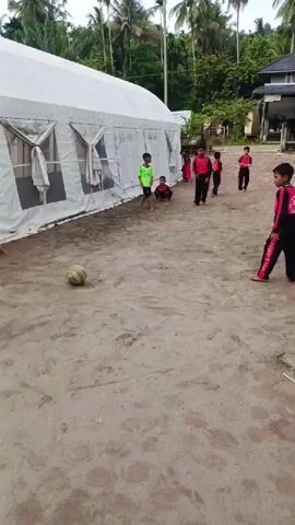 The video shows a group of children playing football (soccer) outdoors in a sandy area. Eight children are visible, most wearing matching black and red tracksuits. One child is wearing a green shirt. They are actively kicking and chasing a white soccer ball. Behind them is a large white tent-like structure with closed side panels. To the right, there's a house with a dark roof, surrounded by green foliage and tall palm trees in the background. There's also a flagpole standing in the corner of the area. The ground is mostly sand, with a paved section on the right side. The overall impression is a casual, spontaneous game being played in a rural or tropical setting.